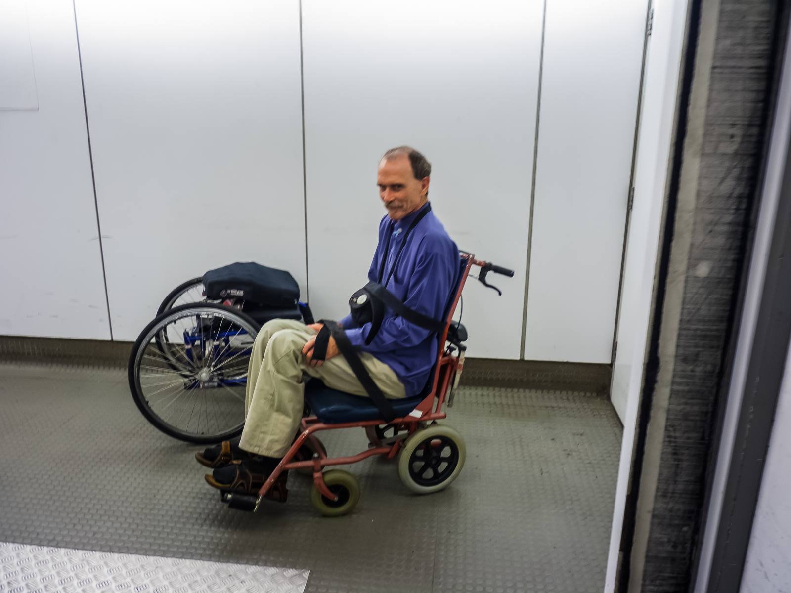 A man seated in a narrow red aisle chair inside an airbridge, with his wheelchair and its large wheels beside him