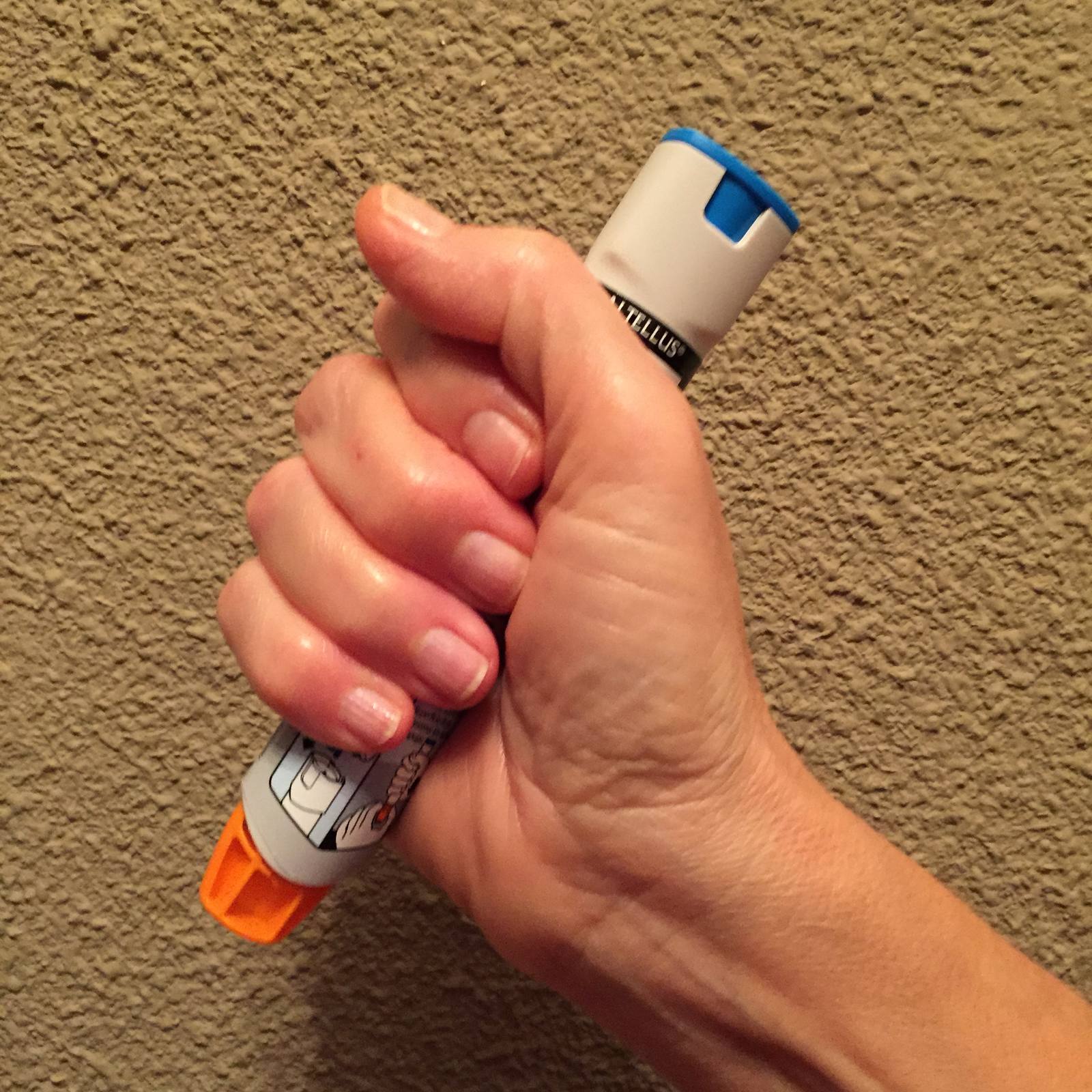 A hand holding an adrenaline auto-injector with a blue safety cap and an orange tip, against a carpeted floor