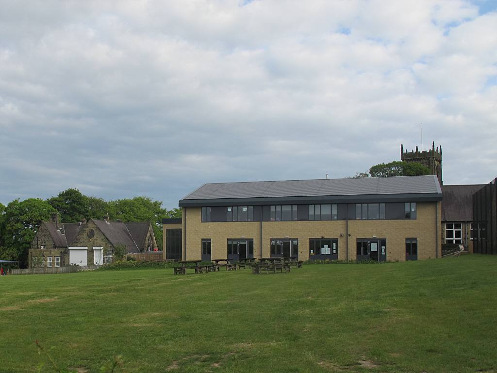 A modern primary school building seen across a grass playing field, with an older school building and a church tower nearby