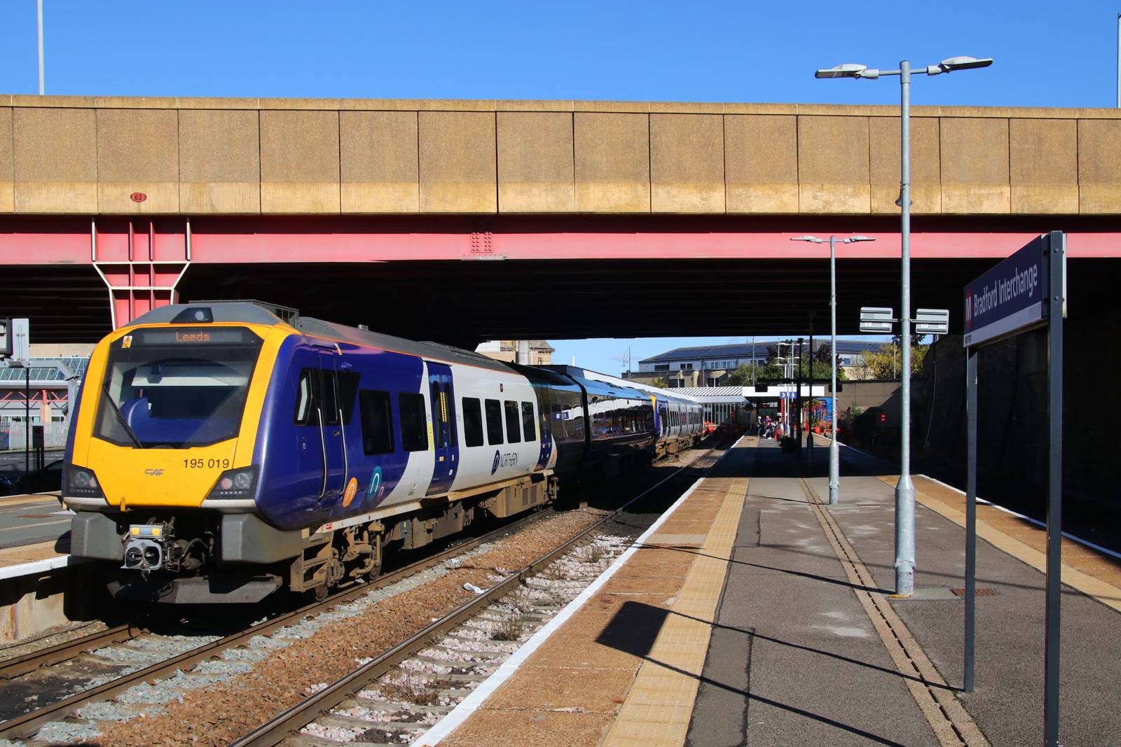 A purple and yellow Northern Class 195 train showing Leeds on its destination display at Bradford Interchange, with the station sign in the foreground