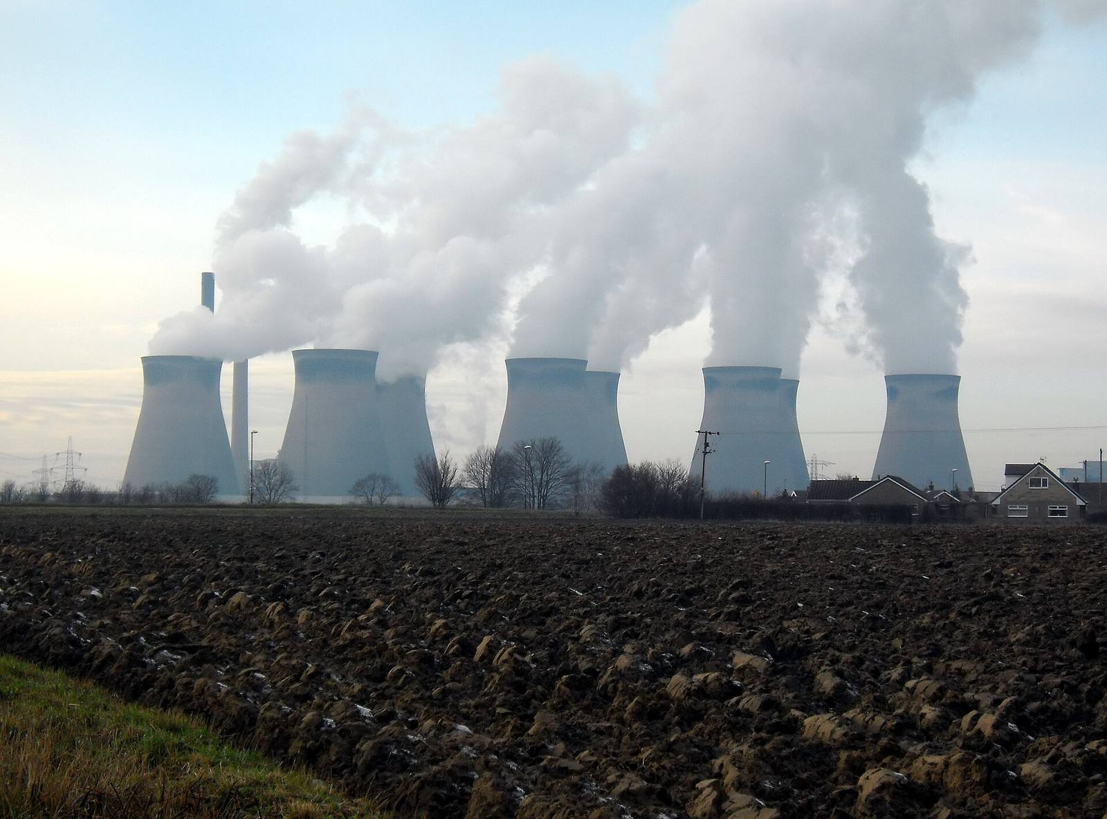 Five power station cooling towers releasing vapour, seen across a ploughed field with houses to one side