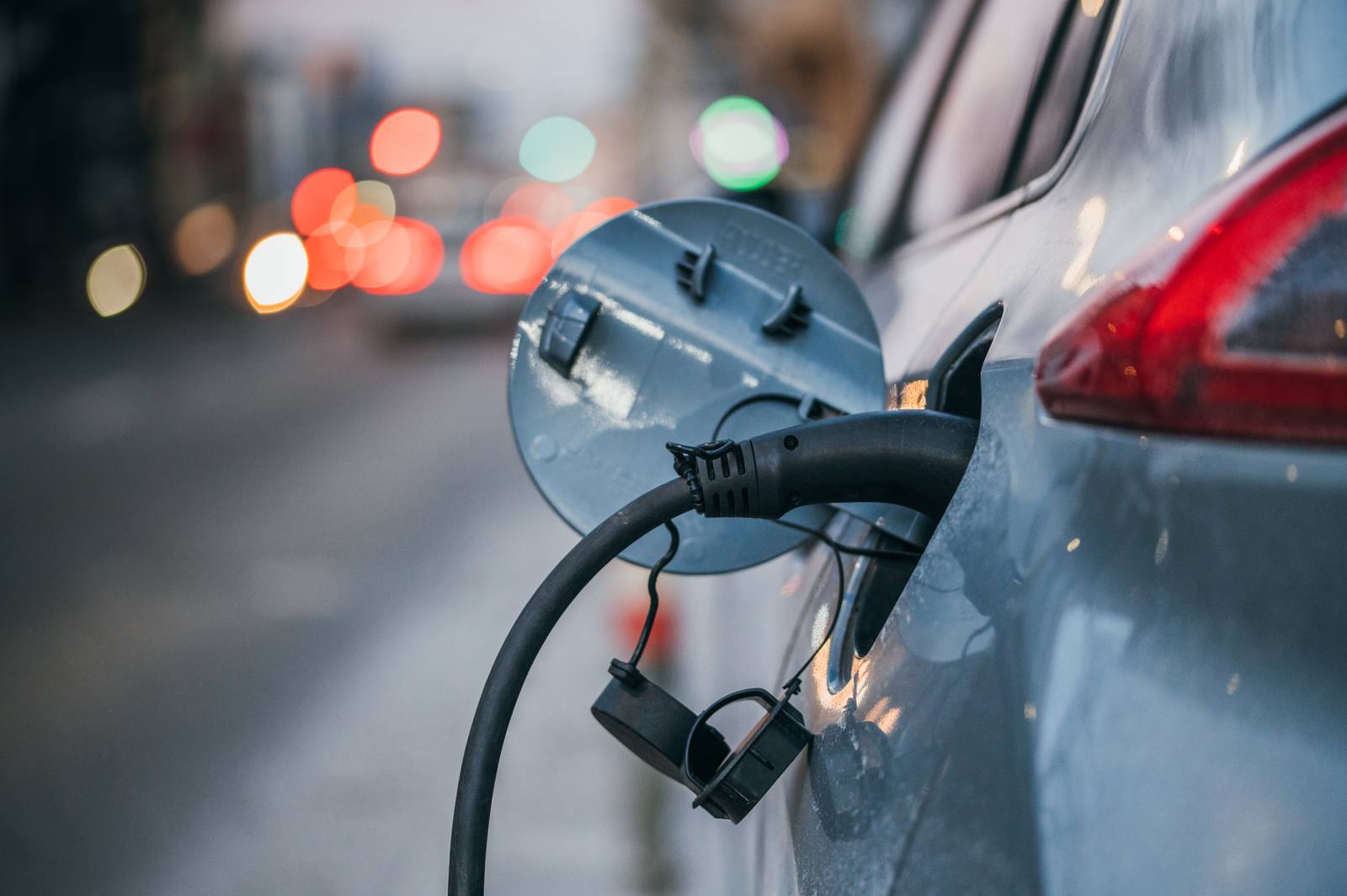 The rear of a blue electric car with a charging cable plugged in, with blurred traffic lights in the background