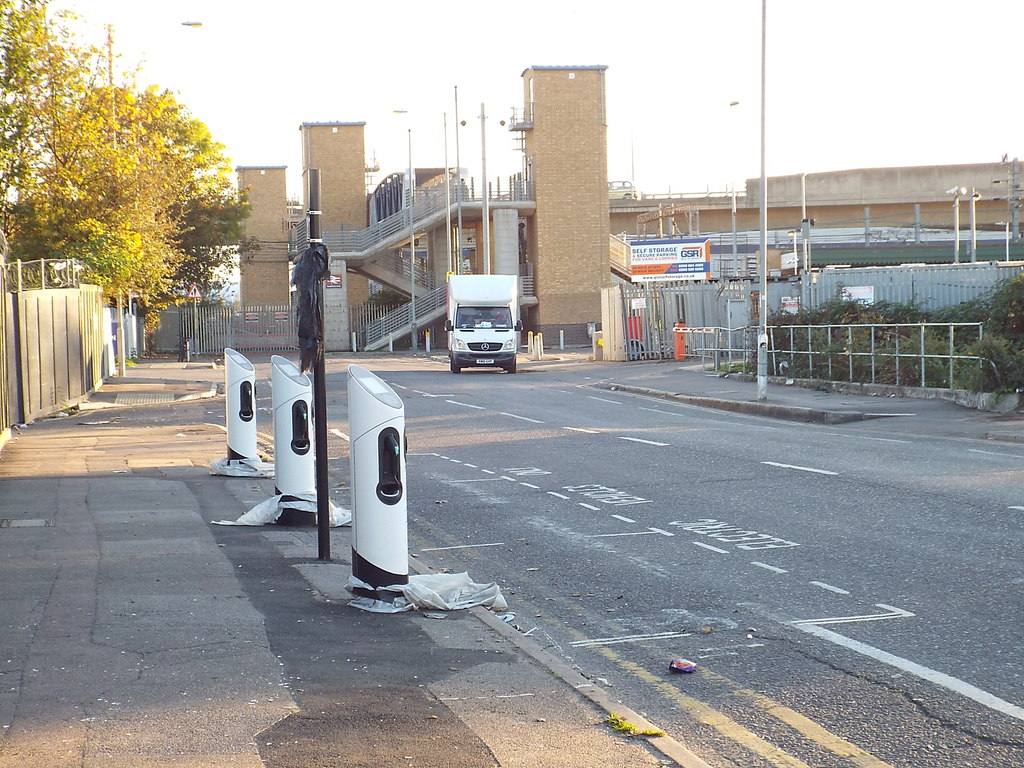 Three white on-street charging bollards standing on a pavement beside a road, with a railway footbridge in the background