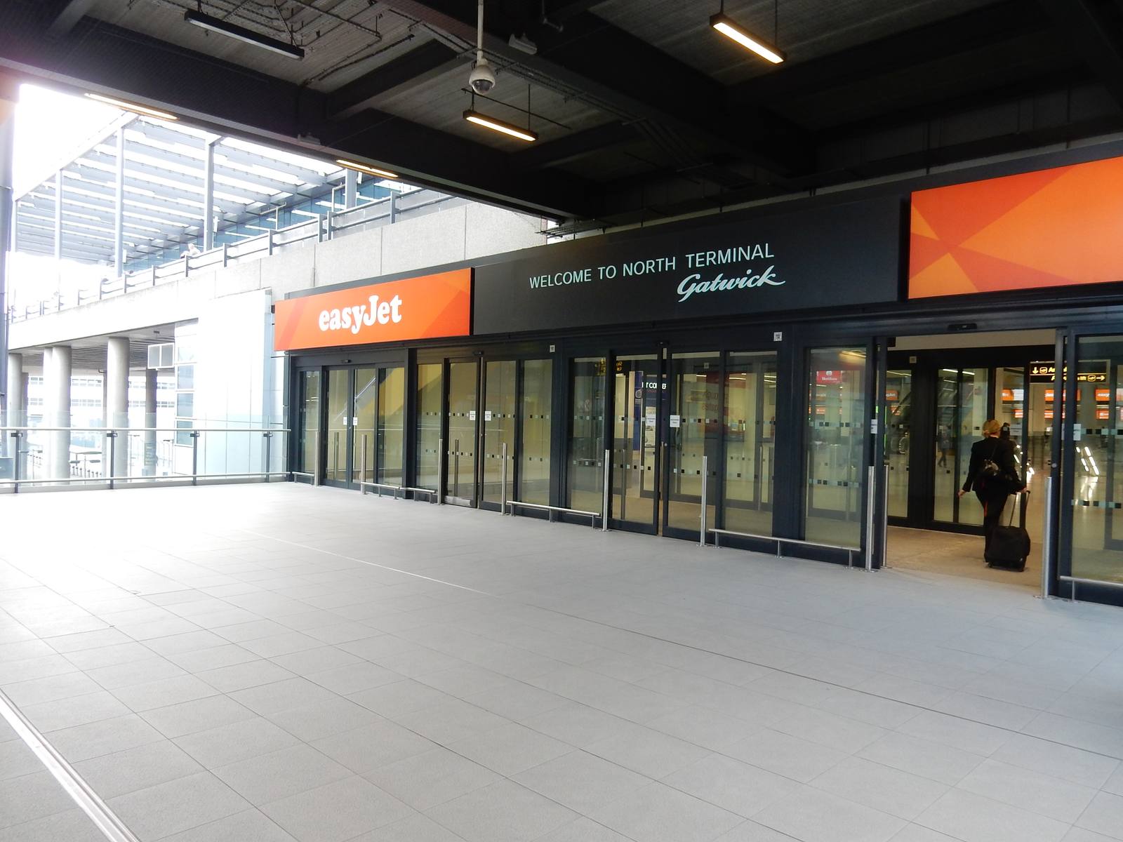 The entrance doors of Gatwick Airport's North Terminal beneath a sign reading Welcome to North Terminal Gatwick, with a passenger wheeling a suitcase inside