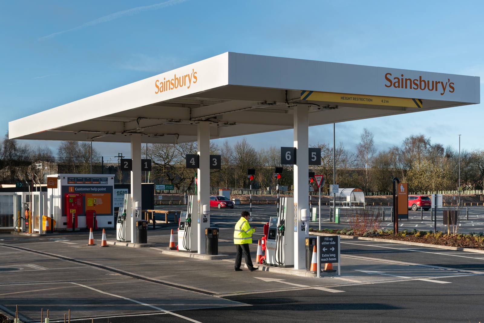 A supermarket petrol station forecourt with rows of fuel pumps under a canopy, and cars parked beyond