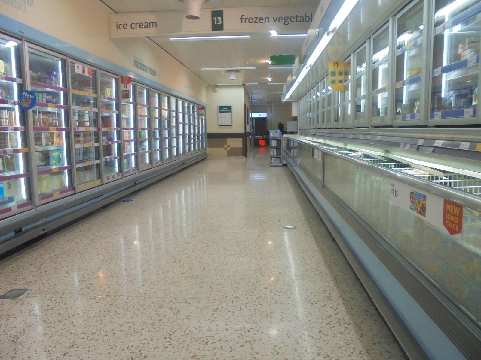 A long supermarket aisle with shelves of tinned and packaged food on both sides and a shopper in the distance