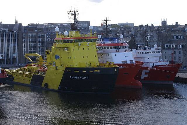 Three offshore support vessels moored in Aberdeen Harbour, a yellow and black vessel in front of two red and white ones, with the city behind