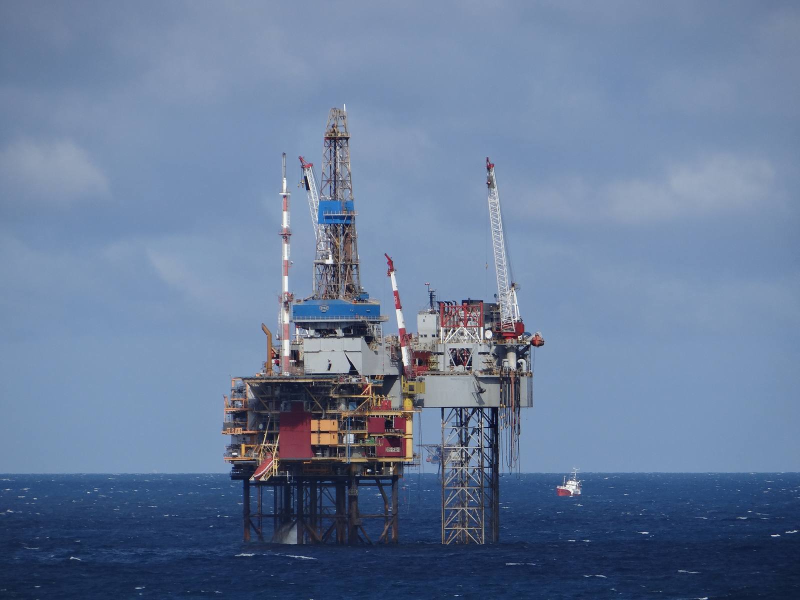 A yellow and red jack-up drilling rig with a tall lattice leg berthed in a harbour, with cranes behind it