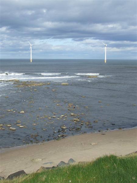 Two offshore wind turbines standing in a grey sea beyond a rocky shore and a sandy beach at Blyth