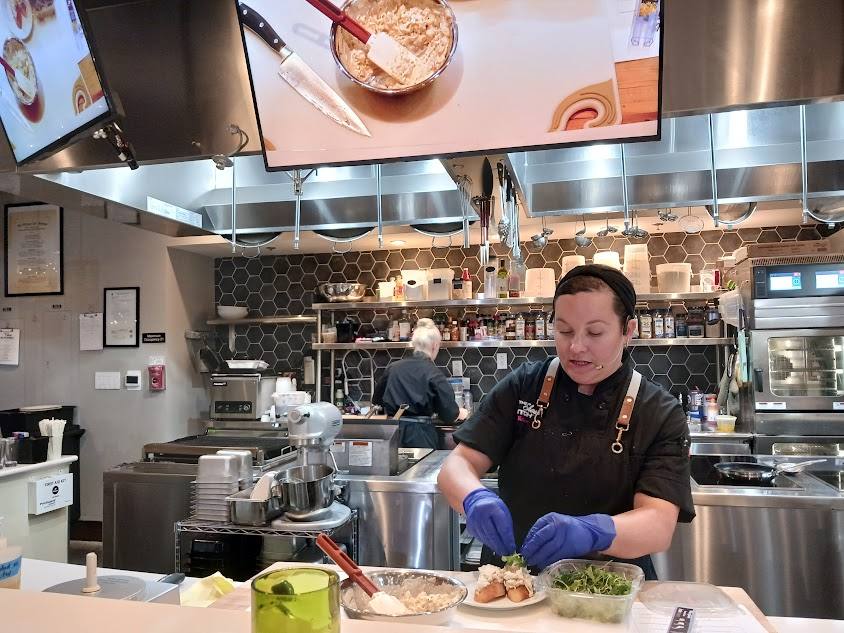 A chef preparing food in a demonstration kitchen, with an overhead screen showing the work surface