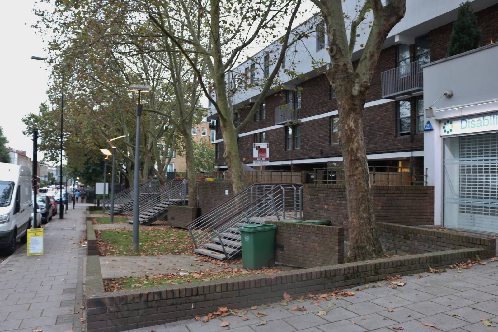 A brick block of council flats behind plane trees on St Pauls Road in Canonbury, with steps and a green bin in the foreground