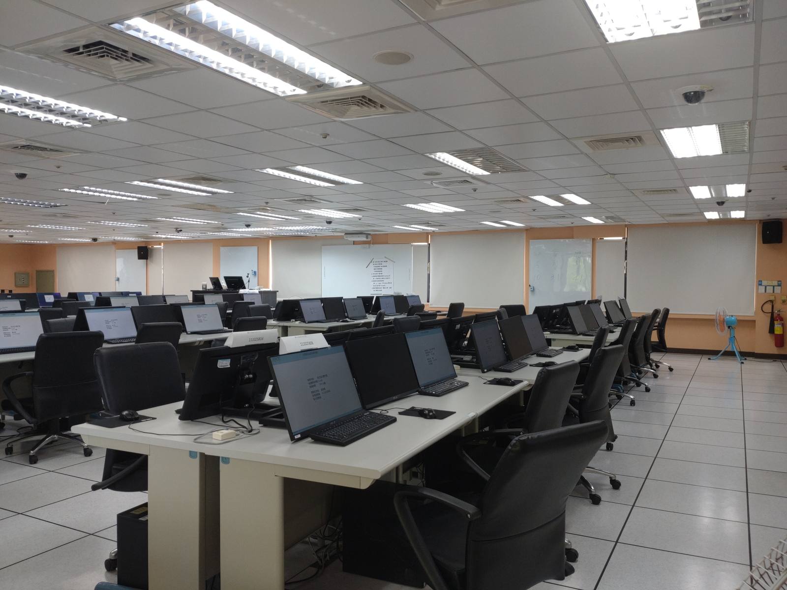 An empty computer classroom with rows of desks carrying laptops and monitors, under strip lighting
