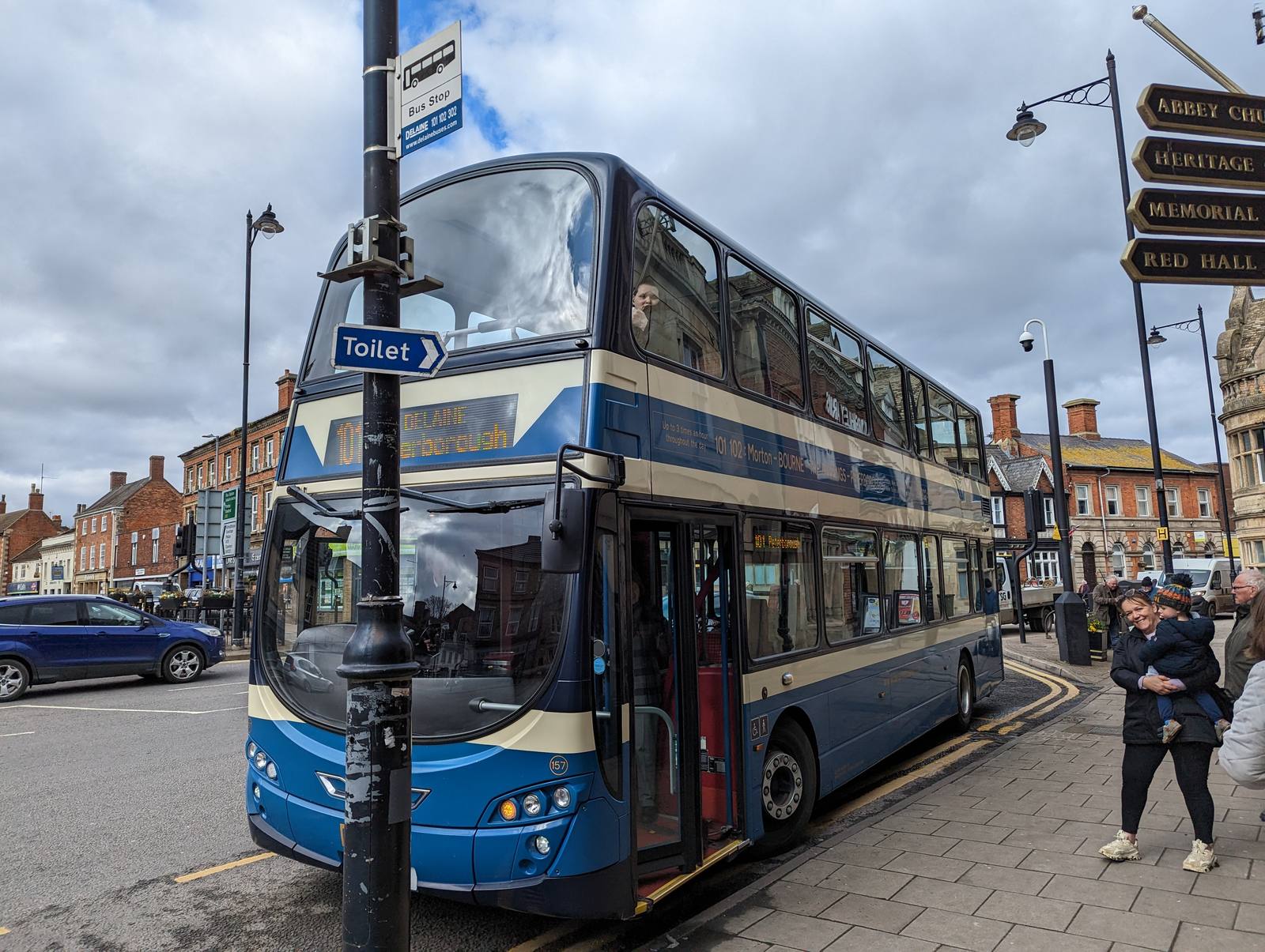 A blue and cream double decker bus stopped at a bus stop sign in a town centre, with pedestrians on the pavement